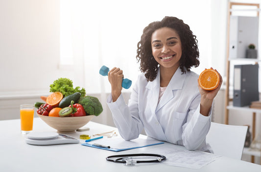 A smiling nutritionist in a white coat holds a dumbbell in one hand and an orange in the other, surrounded by fresh fruits and vegetables on a table, with a notepad and orange juice nearby.