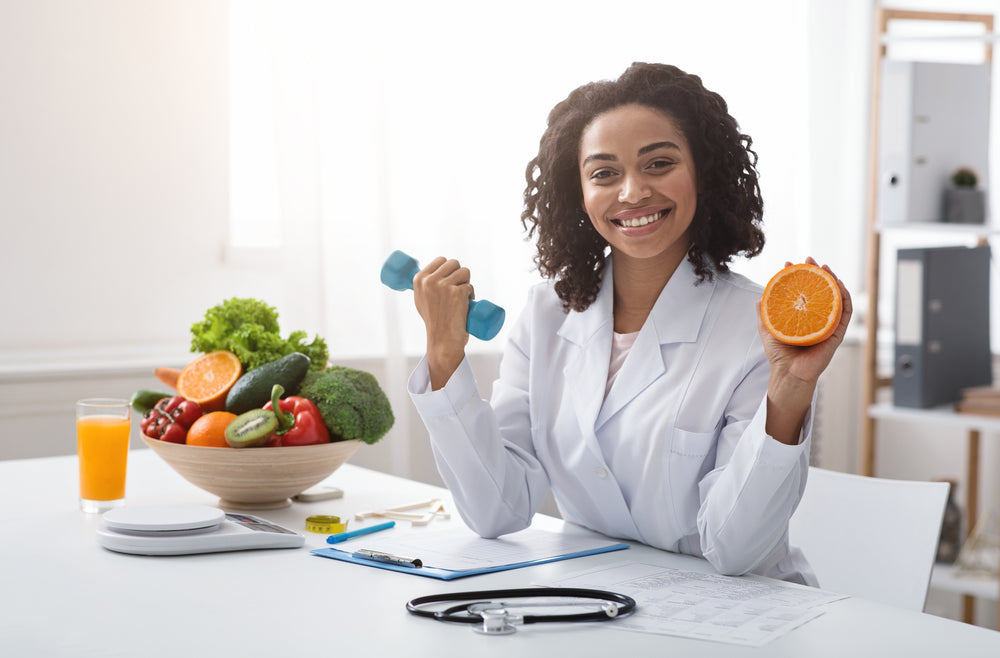 A smiling nutritionist in a white coat holds a dumbbell in one hand and an orange in the other, surrounded by fresh fruits and vegetables on a table, with a notepad and orange juice nearby.
