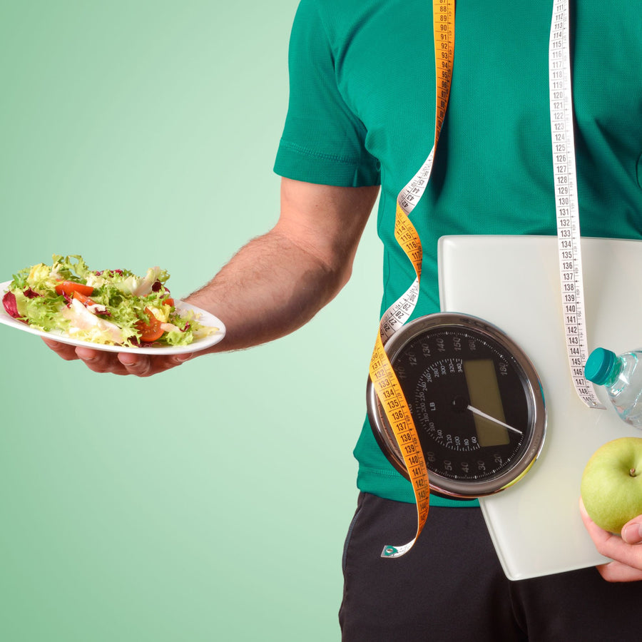 A person in a green shirt holds a plate of salad in one hand and a green apple in the other, while a scale and measuring tape are visible, suggesting a focus on healthy eating and weight loss.