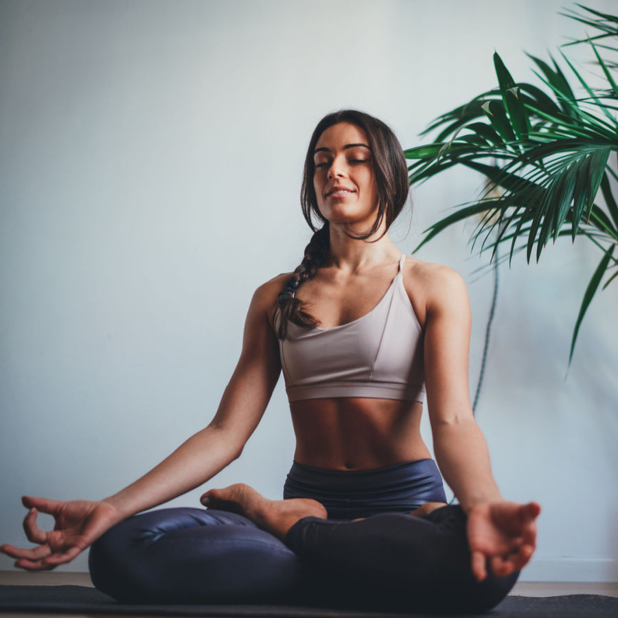 A woman sits cross-legged on a yoga mat, eyes closed and smiling gently, practicing meditation in a bright room with a green plant in the background.