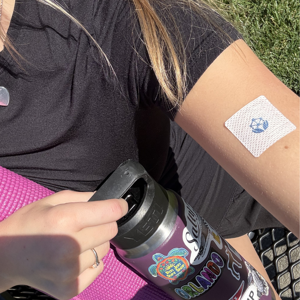 A person in a black shirt sits on a yoga mat, holding a water bottle while a bandage with a blue symbol is visible on their arm, with grass in the background.