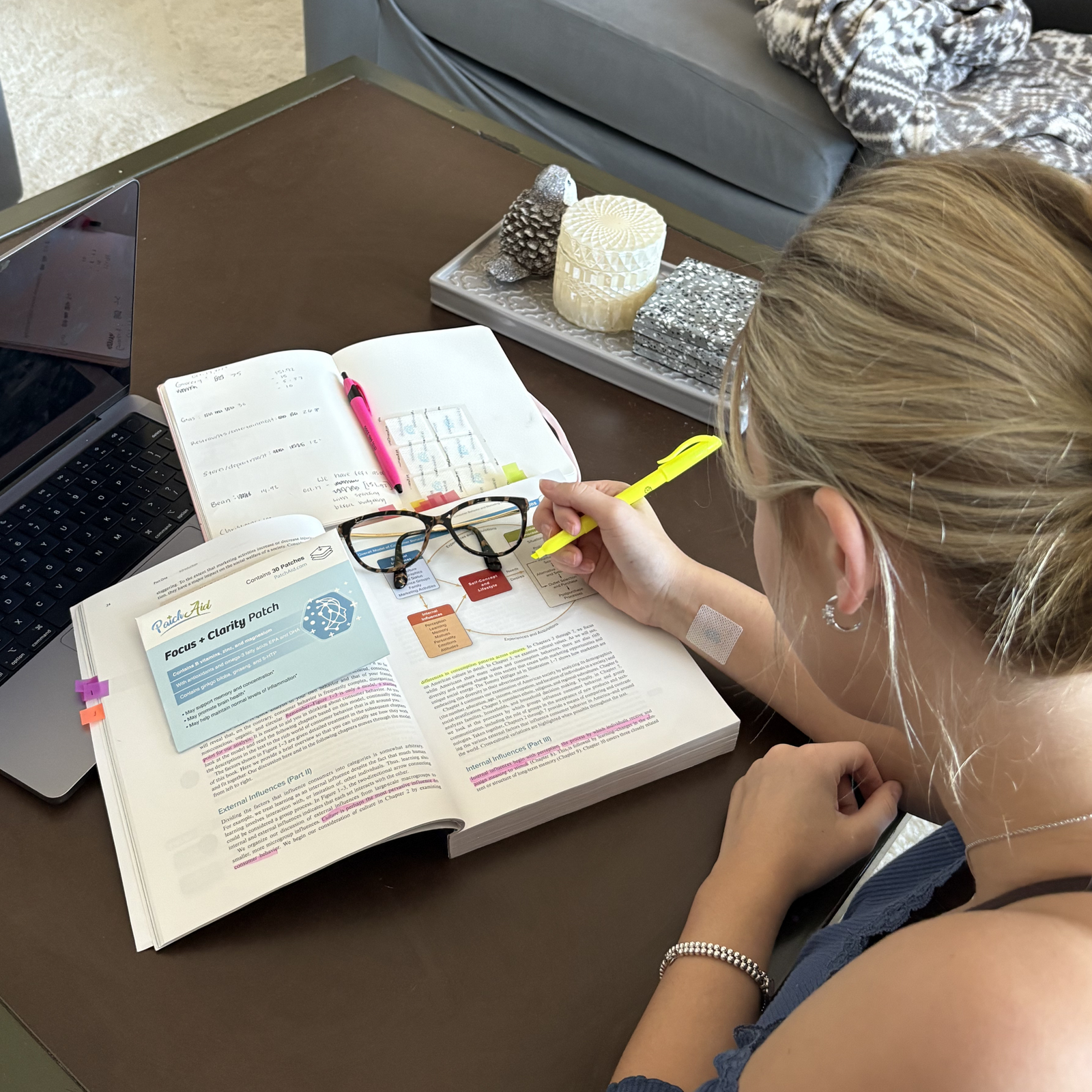 A young woman with blonde hair highlights text in a book while wearing glasses, surrounded by a laptop, stationery, and decorative items on a coffee table in a cozy living room setting.