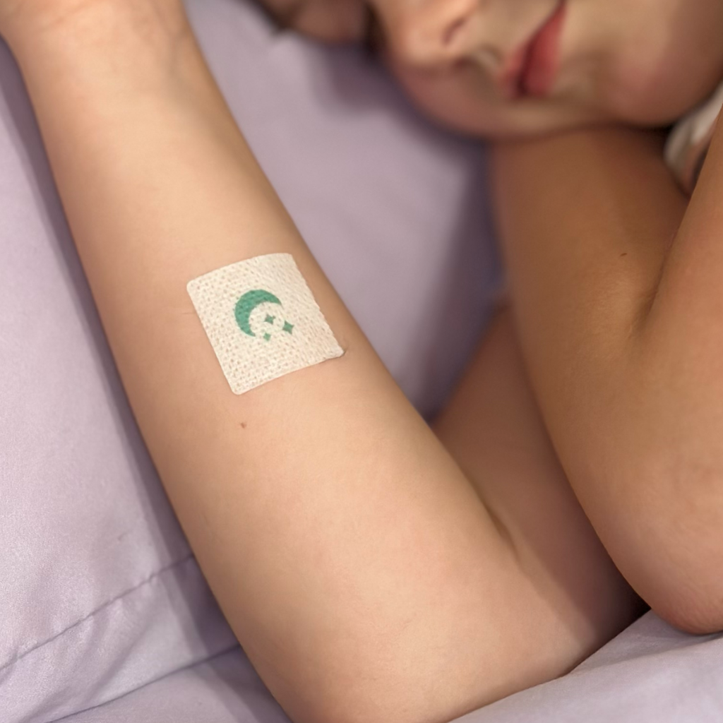 A child is resting with their eyes closed, and a colorful adhesive bandage is placed on their arm, against a soft purple background.