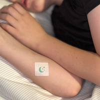 A child is resting on a bed with their arms crossed, displaying a bandage featuring a moon and stars design on one arm.