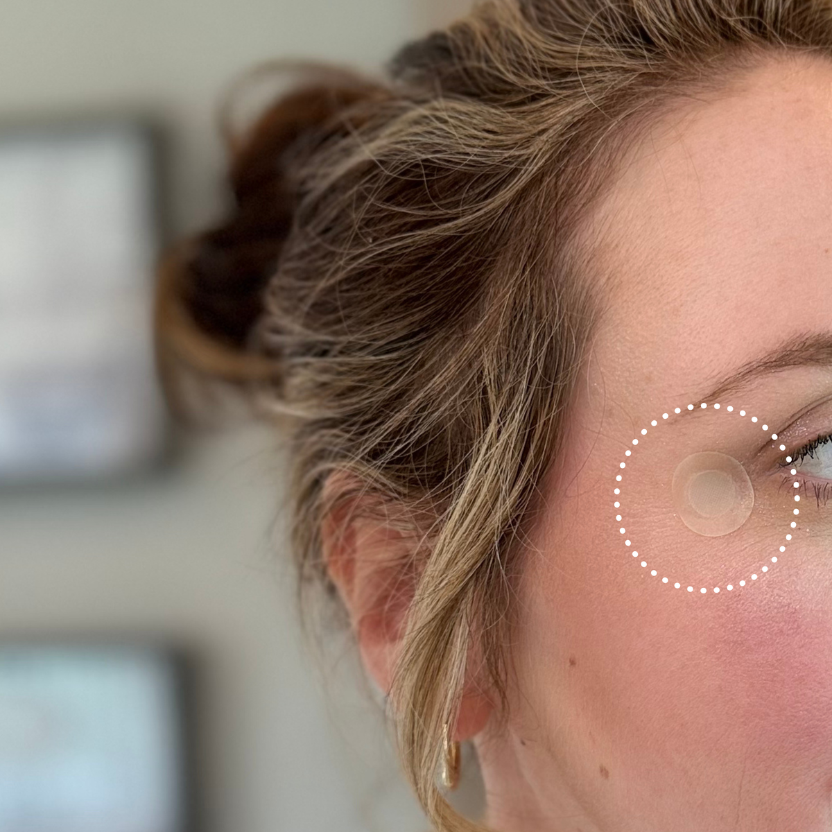 A woman with light brown hair and earrings looks to the side, showcasing a clear skincare patch applied near her eye against a softly blurred background.