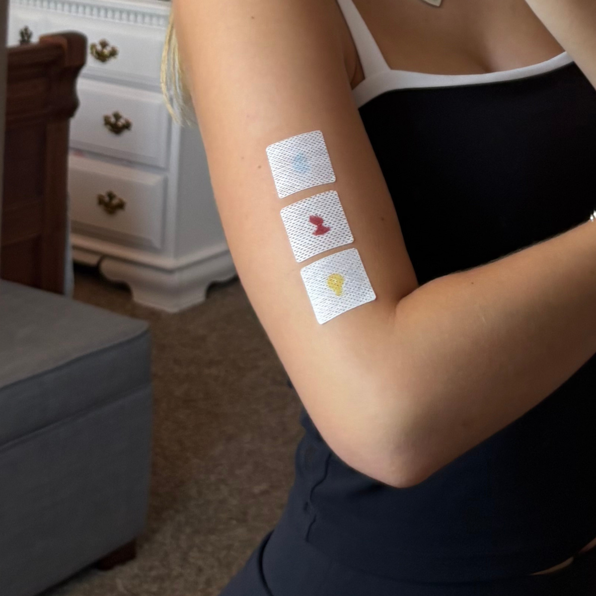 A person with long hair is showing their arm, which has three colorful adhesive patches in a row, against a backdrop of a cozy room with a gray couch and a white dresser.